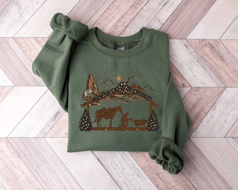 Puede incluir: Una sudadera verde con un gr&aacute;fico marr&oacute;n que representa una escena de la Natividad, con una estrella, monta&ntilde;as y &aacute;rboles. El dise&ntilde;o incluye un caballo, una persona arrodillada y un beb&eacute; en un pesebre. Las mangas est&aacute;n dobladas.