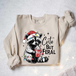 May include: A beige sweatshirt with a cute raccoon wearing a Santa hat and holding a cup of hot cocoa. The raccoon is surrounded by Christmas lights. The text "Cute But Feral" is printed on the sweatshirt.