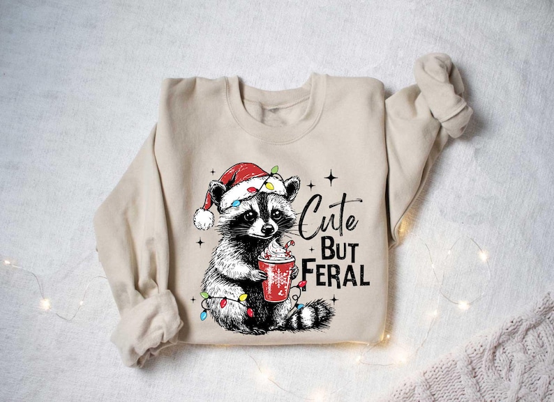 May include: A beige sweatshirt with a cute raccoon wearing a Santa hat and holding a cup of hot cocoa. The raccoon is surrounded by Christmas lights. The text "Cute But Feral" is printed on the sweatshirt.