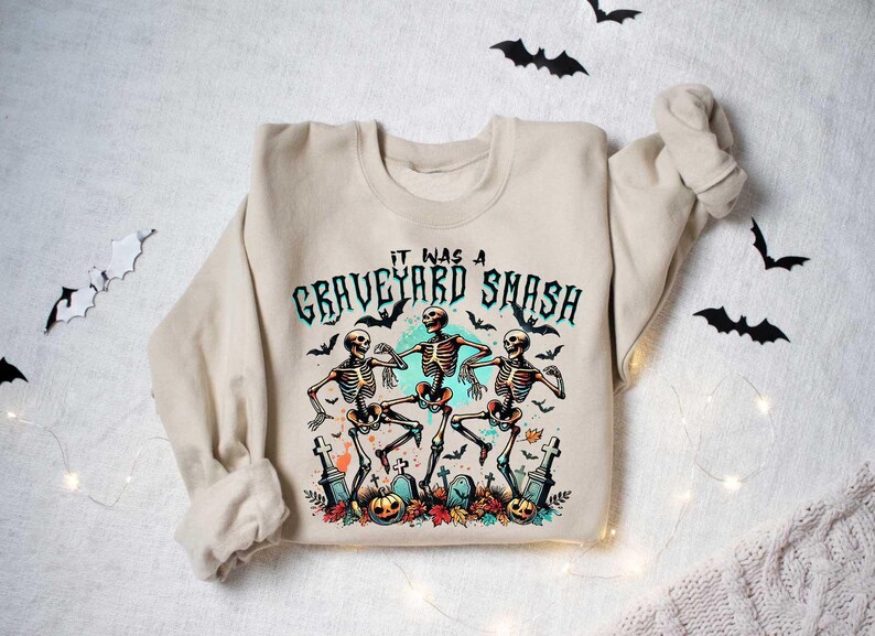 May include: A beige sweatshirt with a graphic print of three skeletons dancing in a graveyard. The text "It was a Graveyard Smash" is printed above the skeletons.