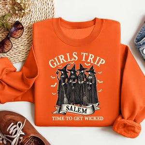 May include: Orange sweatshirt with a graphic of three witches in black dresses and pointy hats. The text "Girls Trip Salem Time to Get Wicked" is printed on the sweatshirt.