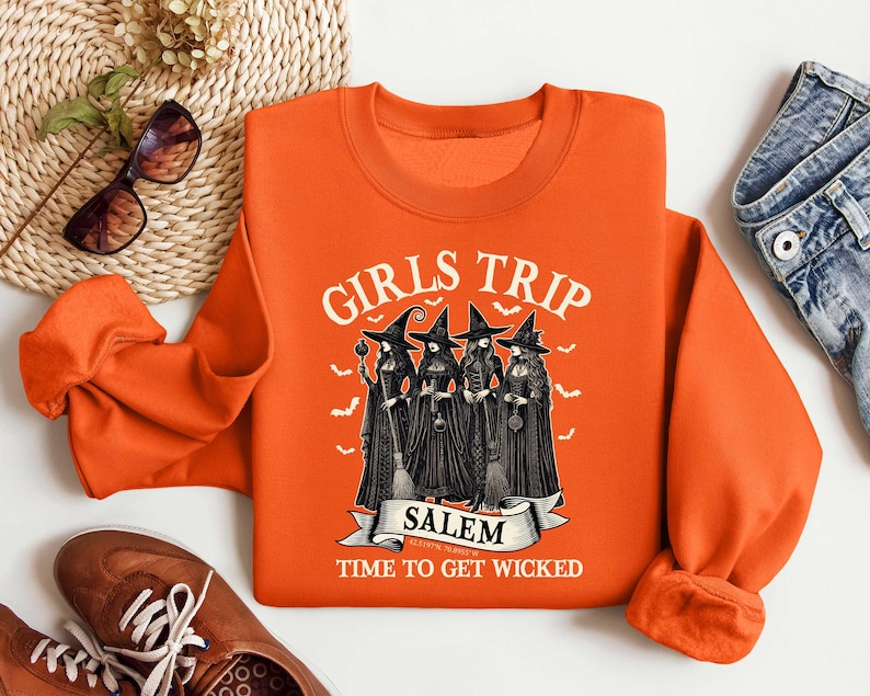 May include: Orange sweatshirt with a graphic of three witches in black dresses and pointy hats. The text "Girls Trip Salem Time to Get Wicked" is printed on the sweatshirt.