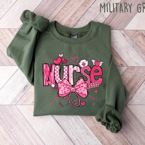Peut inclure: Un sweat-shirt vert avec le mot "Nurse" en lettres roses avec un n&oelig;ud rose et d'autres symboles m&eacute;dicaux. Le sweat-shirt est sur un fond en bois.