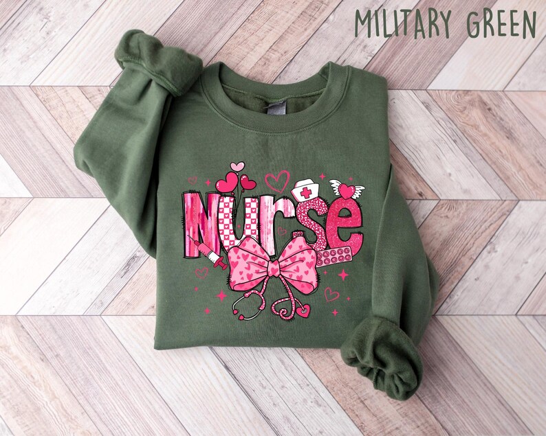 Peut inclure: Un sweat-shirt vert avec le mot "Nurse" en lettres roses avec un n&oelig;ud rose et d'autres symboles m&eacute;dicaux. Le sweat-shirt est sur un fond en bois.