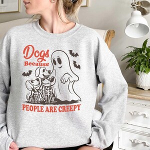Puede incluir: Una sudadera gris con un estampado gr&aacute;fico de un fantasma y tres perros. El texto en la sudadera dice "Dogs Because People Are Creepy."