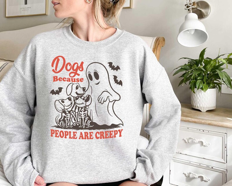Puede incluir: Una sudadera gris con un estampado gr&aacute;fico de un fantasma y tres perros. El texto en la sudadera dice "Dogs Because People Are Creepy."