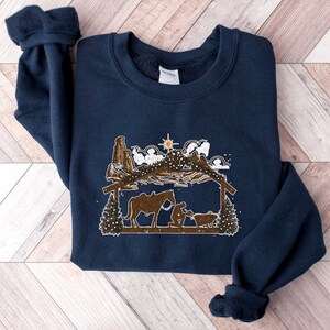 Puede incluir: Sudadera azul marino con un dise&ntilde;o marr&oacute;n y blanco de un pesebre. El dise&ntilde;o incluye un establo, figuras, un caballo y una estrella. La sudadera tiene cuello redondo y mangas largas con pu&ntilde;os.