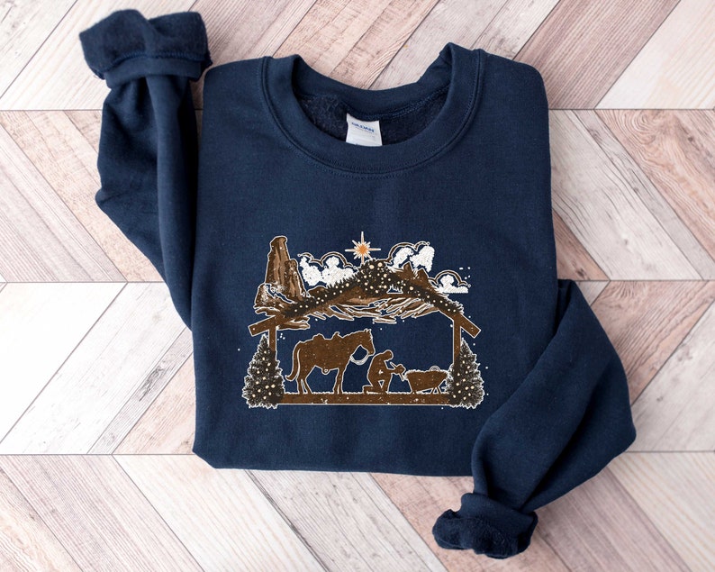 Puede incluir: Sudadera azul marino con un dise&ntilde;o marr&oacute;n y blanco de un pesebre. El dise&ntilde;o incluye un establo, figuras, un caballo y una estrella. La sudadera tiene cuello redondo y mangas largas con pu&ntilde;os.