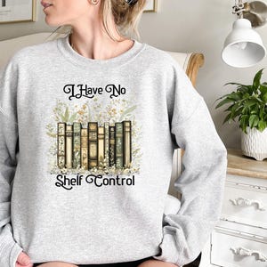 K&ouml;nnte beinhalten: Ein graues Sweatshirt mit dem Text "I Have No Shelf Control" auf der Vorderseite. Der Text ist von einem floralen Design und einer Illustration eines B&uuml;cherstapels umgeben.