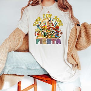 Pode incluir: Uma camiseta creme com um gr&aacute;fico colorido com o texto "TIME FOR SOME FIESTA". O gr&aacute;fico inclui personagens de desenhos animados em trajes festivos. A camisa &eacute; de manga curta e tem gola redonda.
