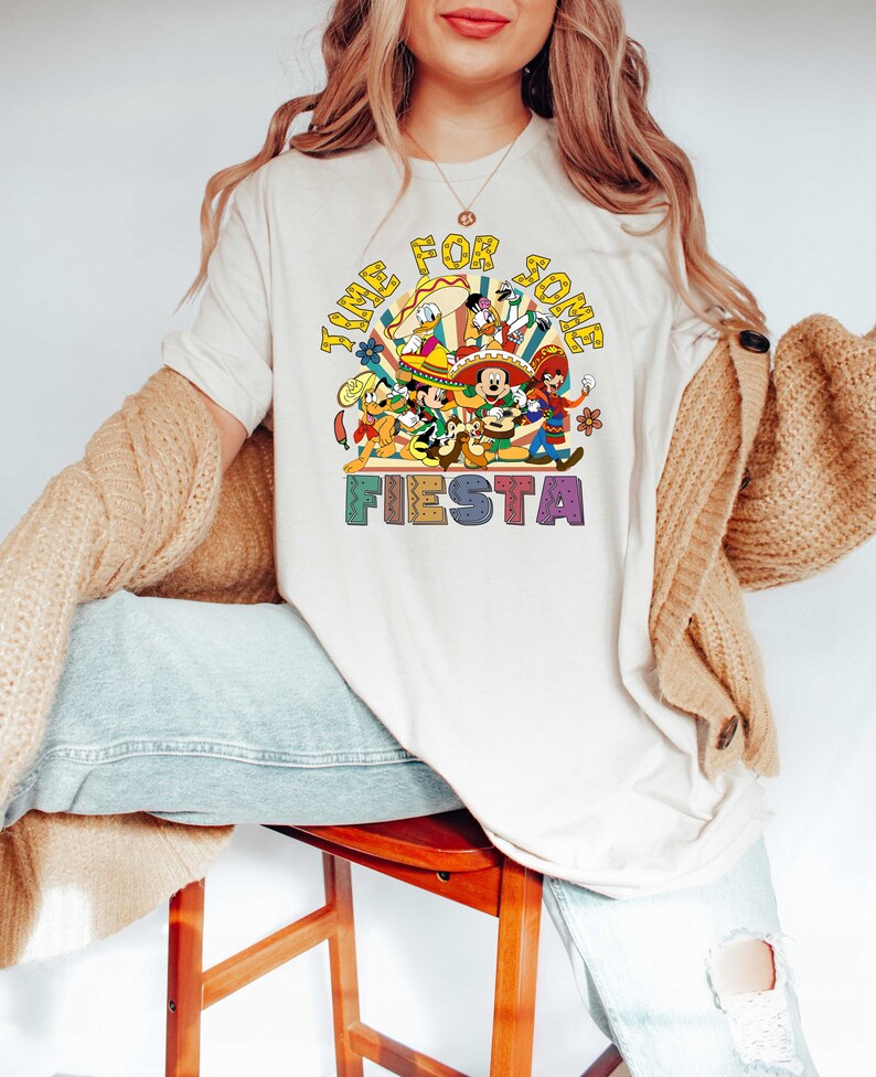 Pode incluir: Uma camiseta creme com um gr&aacute;fico colorido com o texto "TIME FOR SOME FIESTA". O gr&aacute;fico inclui personagens de desenhos animados em trajes festivos. A camisa &eacute; de manga curta e tem gola redonda.