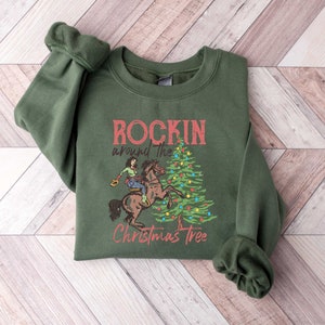 May include: Green sweatshirt with a Christmas-themed graphic. The graphic features a cowboy riding a horse around a decorated Christmas tree. The text reads "Rockin' around the Christmas Tree".