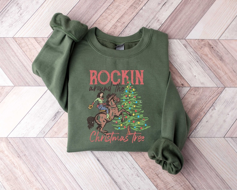 May include: Green sweatshirt with a Christmas-themed graphic. The graphic features a cowboy riding a horse around a decorated Christmas tree. The text reads "Rockin' around the Christmas Tree".