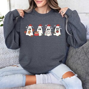 Op de afbeelding: Een donkergrijs sweatshirt met een feestelijk ontwerp met vier geesten die kerstmanmutsen dragen. De geesten houden een peperkoekman, een cadeau, een lichtsnoer en een snoepstok.