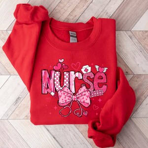 Peut inclure: Sweat-shirt rouge avec un motif rose et blanc qui dit "Infirmi&egrave;re" avec des c&oelig;urs et d'autres symboles m&eacute;dicaux.