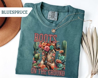 May include: Teal Comfort Colors t-shirt with a graphic of cowboy boots, cacti, and flowers. The text "BOOTS ON THE GROUND" is displayed in a red, Western-style font. A gold necklace is draped across the shirt.