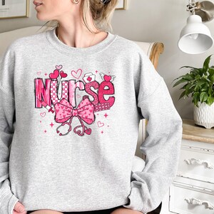 Peut inclure: Sweat-shirt gris avec un motif graphique rose qui dit "Nurse" avec des c&oelig;urs, un n&oelig;ud et un st&eacute;thoscope.
