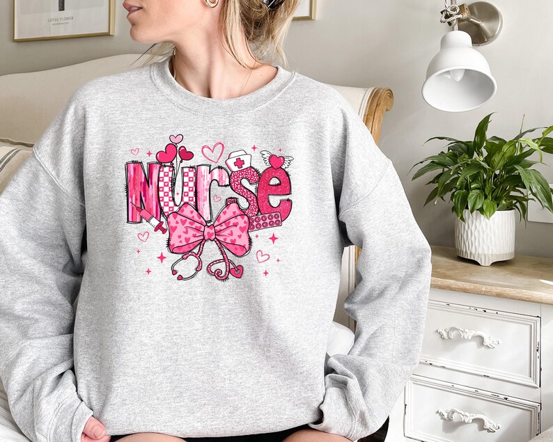 Peut inclure: Sweat-shirt gris avec un motif graphique rose qui dit "Nurse" avec des c&oelig;urs, un n&oelig;ud et un st&eacute;thoscope.