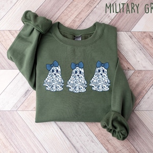 Może przedstawiać: Zielona bluza w kolorze khaki z nadrukiem trzech niebieskich i białych duch&oacute;w z niebieskimi kokardkami. Tekst "MILITARY GREEN" jest nadrukowany na g&oacute;rze obrazu na biało.