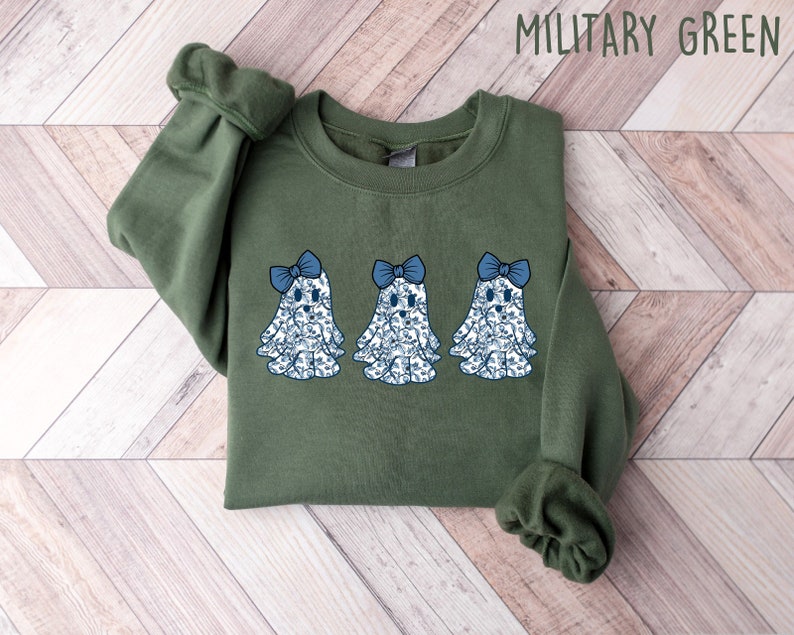 Może przedstawiać: Zielona bluza w kolorze khaki z nadrukiem trzech niebieskich i białych duch&oacute;w z niebieskimi kokardkami. Tekst "MILITARY GREEN" jest nadrukowany na g&oacute;rze obrazu na biało.