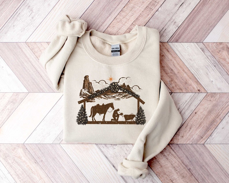 Puede incluir: Sudadera de color crema con un gr&aacute;fico marr&oacute;n de una escena de la natividad con un caballo, una figura arrodillada y una estrella. El dise&ntilde;o incluye una cordillera y una estructura de madera, evocando un tema r&uacute;stico y occidental. La sudadera tiene cuello redondo y mangas largas.