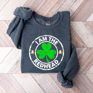Puede incluir: Una sudadera gris oscuro con un dise&ntilde;o circular blanco que dice "I am the Redhead" con un tr&eacute;bol verde dentro del c&iacute;rculo.