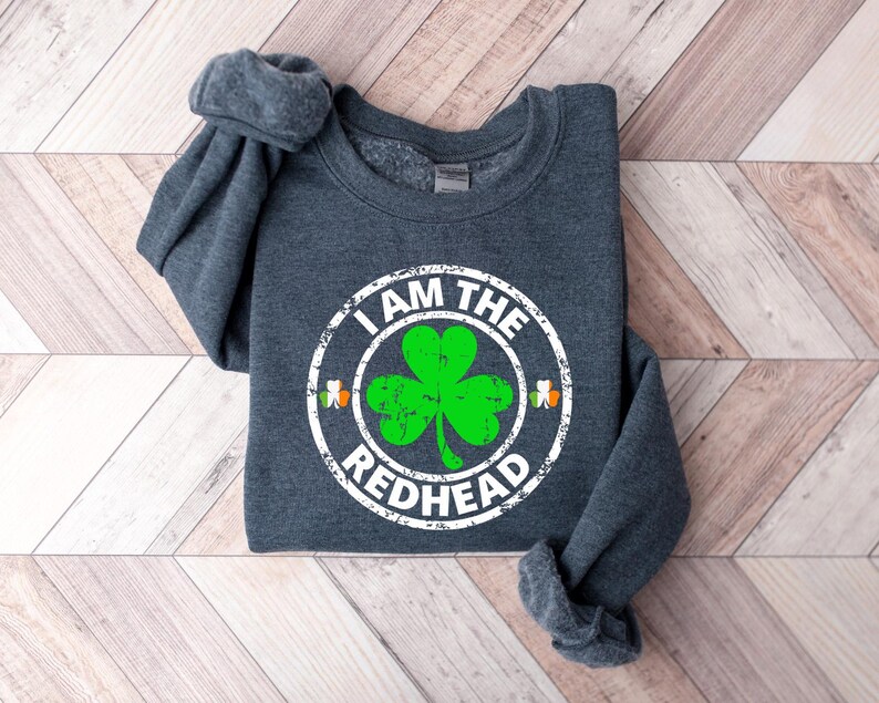 Puede incluir: Una sudadera gris oscuro con un dise&ntilde;o circular blanco que dice "I am the Redhead" con un tr&eacute;bol verde dentro del c&iacute;rculo.