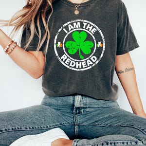 Puede incluir: Una camiseta gris oscuro con un gr&aacute;fico circular blanco desgastado que presenta un tr&eacute;bol verde y el texto "I AM THE REDHEAD".