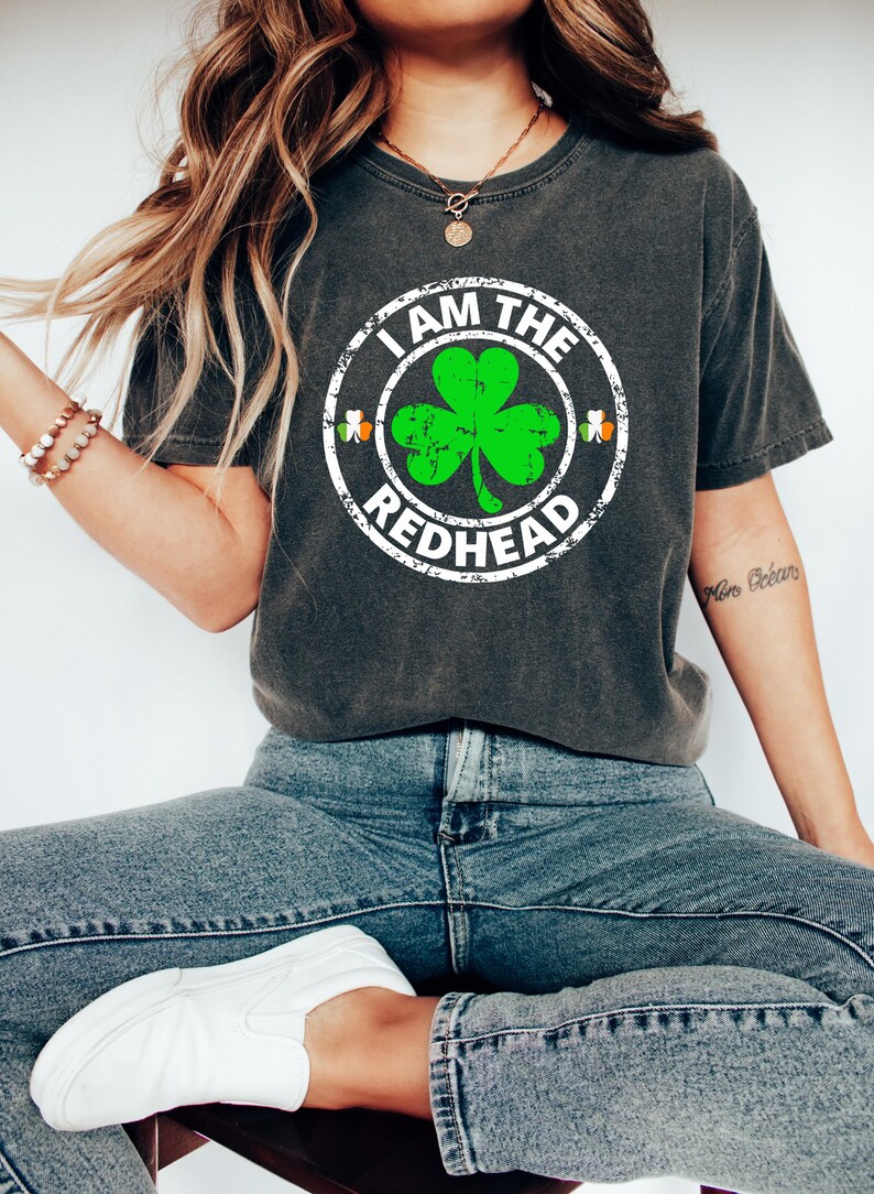 Puede incluir: Una camiseta gris oscuro con un gr&aacute;fico circular blanco desgastado que presenta un tr&eacute;bol verde y el texto "I AM THE REDHEAD".