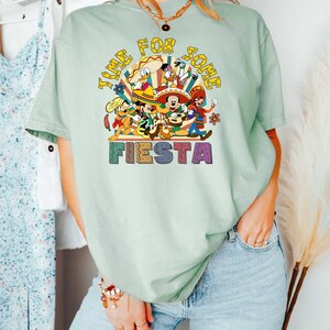 Pode incluir: Camiseta verde claro com um gr&aacute;fico colorido que diz "Time for some Fiesta". O gr&aacute;fico apresenta personagens de desenhos animados em trajes festivos. A camisa &eacute; usada com jeans azuis.