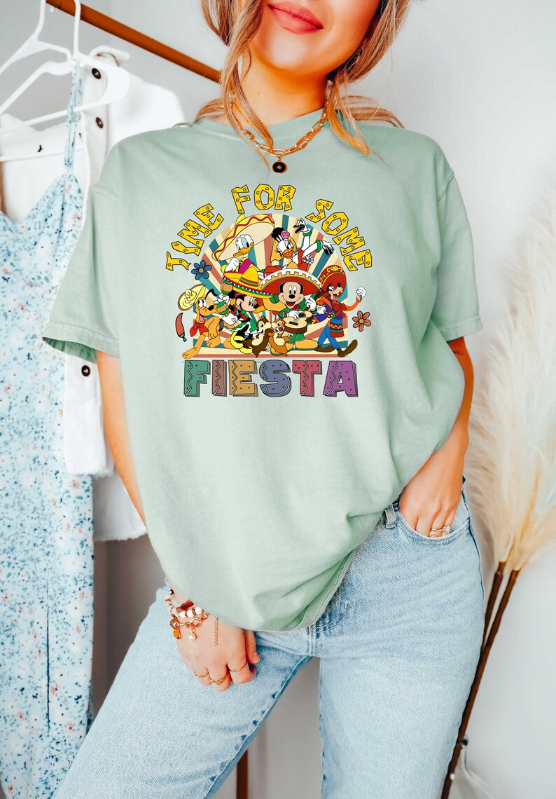 Pode incluir: Camiseta verde claro com um gr&aacute;fico colorido que diz "Time for some Fiesta". O gr&aacute;fico apresenta personagens de desenhos animados em trajes festivos. A camisa &eacute; usada com jeans azuis.