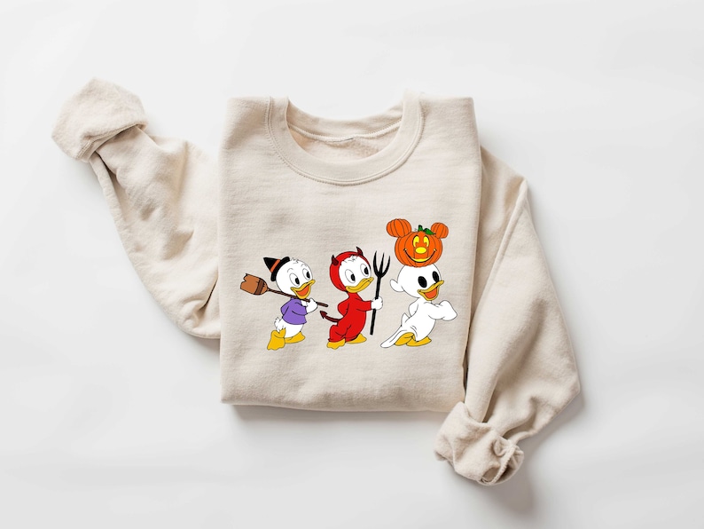 May include: A beige sweatshirt featuring a printed design of three cartoon ducks in Halloween costumes. The ducks are dressed as a witch, a devil, and a ghost with a pumpkin on its head.