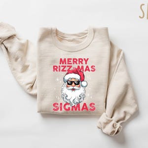 May include: A sand-colored sweatshirt with the text "MERRY RIZZ-MAS SIGMAS" in red. The graphic features Santa Claus wearing sunglasses and a red hat. The sweatshirt is folded with the sleeves rolled up.