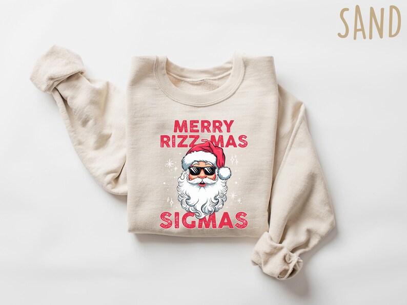 May include: A sand-colored sweatshirt with the text "MERRY RIZZ-MAS SIGMAS" in red. The graphic features Santa Claus wearing sunglasses and a red hat. The sweatshirt is folded with the sleeves rolled up.