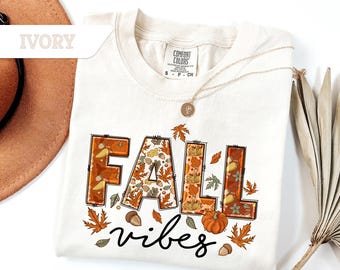 May include: Ivory-colored t-shirt with the words "FALL vibes" in a fall-themed design. The word "FALL" is in orange block letters with autumn leaf and acorn details. A pumpkin and leaves surround the word "vibes".