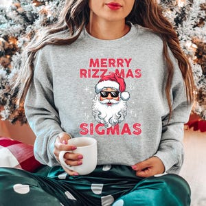 May include: A gray sweatshirt with the text "MERRY RIZZ-MAS SIGMAS" in red, featuring a Santa Claus graphic wearing sunglasses. The sweatshirt is part of a holiday-themed clothing collection.