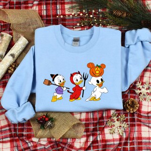 May include: A light blue sweatshirt featuring a graphic print of three cartoon ducks dressed in Halloween costumes. The ducks are wearing a witch hat, devil horns, and a ghost costume with a pumpkin on its head.