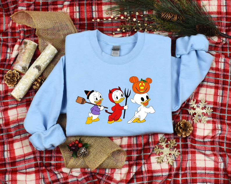 May include: A light blue sweatshirt featuring a graphic print of three cartoon ducks dressed in Halloween costumes. The ducks are wearing a witch hat, devil horns, and a ghost costume with a pumpkin on its head.