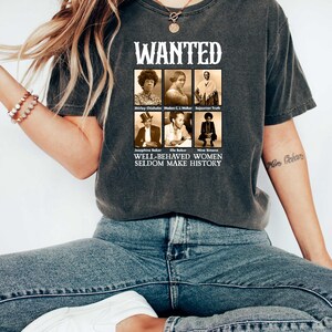 May include: A dark grey t-shirt with a vintage-style graphic featuring portraits of influential women. The graphic includes the text "WANTED" and "WELL-BEHAVED WOMEN SELDOM MAKE HISTORY".