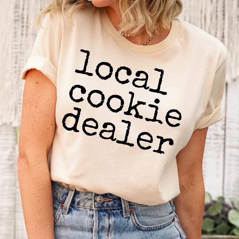 May include: A cream colored t-shirt with the text "local cookie dealer" printed in black.
