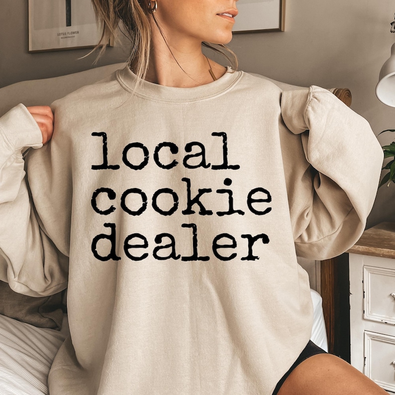 May include: A beige sweatshirt with the text "local cookie dealer" printed in black.