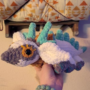May include: A crocheted plush toy dragon with a white body, grey head, orange eyes, and teal spikes. The dragon is being held in a hand with a turquoise and pink beaded bracelet.