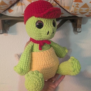 May include: A green crocheted turtle wearing a red crocheted hat and a red scarf. The turtle has large, black eyes and a yellow crocheted shell.