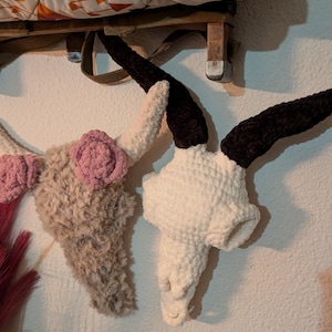 May include: Two crocheted animal skull decorations. One is white with black horns and the other is brown with pink flowers.