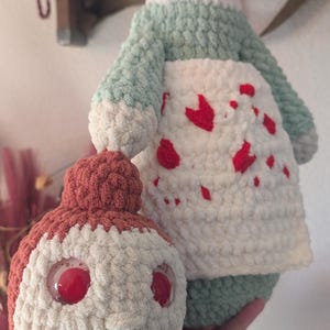 May include: A crocheted plush toy with a red pom-pom hat, white body with red hearts, and a green bodice. The toy is holding a smaller crocheted plush toy with a red and white body and red eyes.