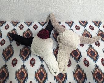 May include: Two crocheted animal skull ornaments, one with black horns and a burgundy flower, the other with beige horns, on a white and brown patterned fabric.