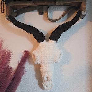 May include: A white crocheted animal skull with black crocheted horns hanging on a wall. The skull is a decorative piece with a rustic aesthetic.