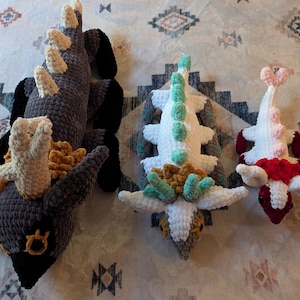 May include: Three crocheted plush toys shaped like dragons. The largest dragon is brown and tan with antlers. The middle dragon is white with green and tan accents. The smallest dragon is white with pink and red accents.