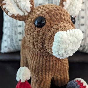 May include: A brown crocheted reindeer with white accents on the ears and nose. The reindeer is wearing a red crocheted Santa hat and a gray and red crocheted scarf.