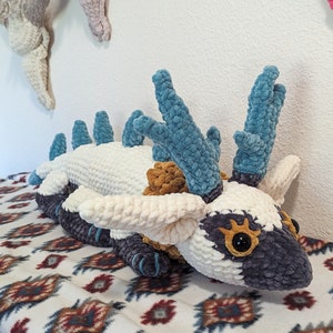 May include: A crocheted plush toy in the shape of a white and gray creature with blue horns. The creature has large, black eyes and a small, orange nose. The toy is lying on a white and brown patterned blanket.